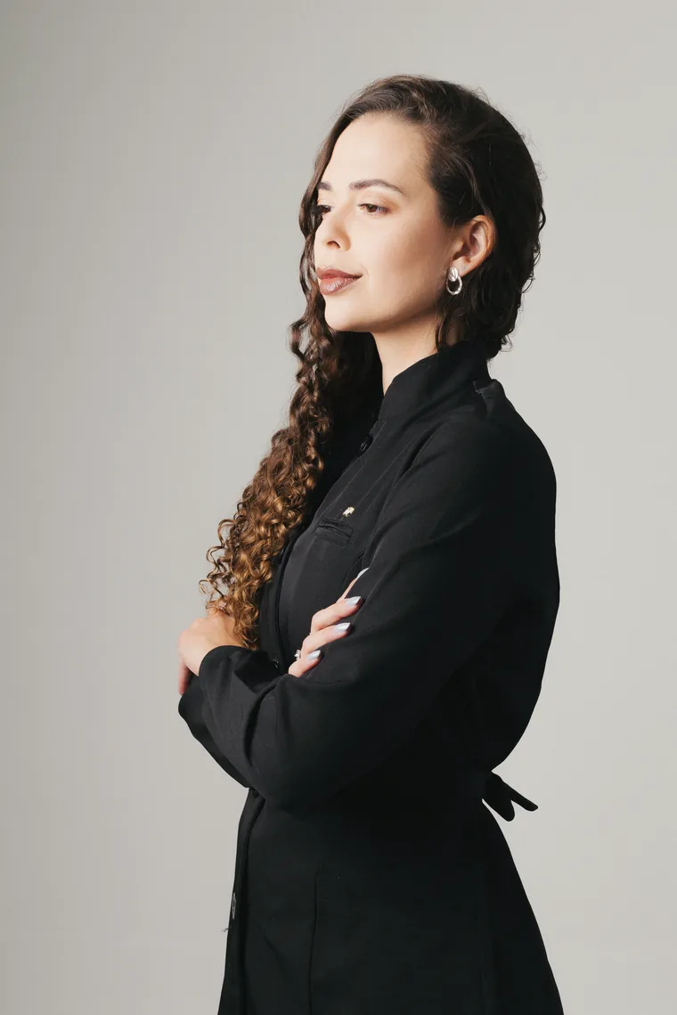 Amanda Neves de perfil, com jaleco preto e braços cruzados, em fundo cinza claro.