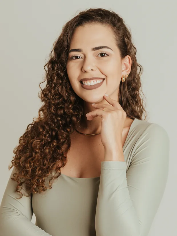 Amanda Neves sorrindo, de blusa verde-oliva, apoiando a mão no queixo.